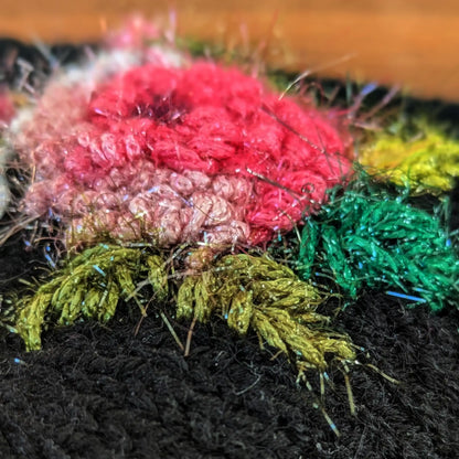 Très gros plan de la rose crochetée puis brodée sur une paire de mitaines noires. La rose est entourée de tiges allant du vert olive au vert kaki.