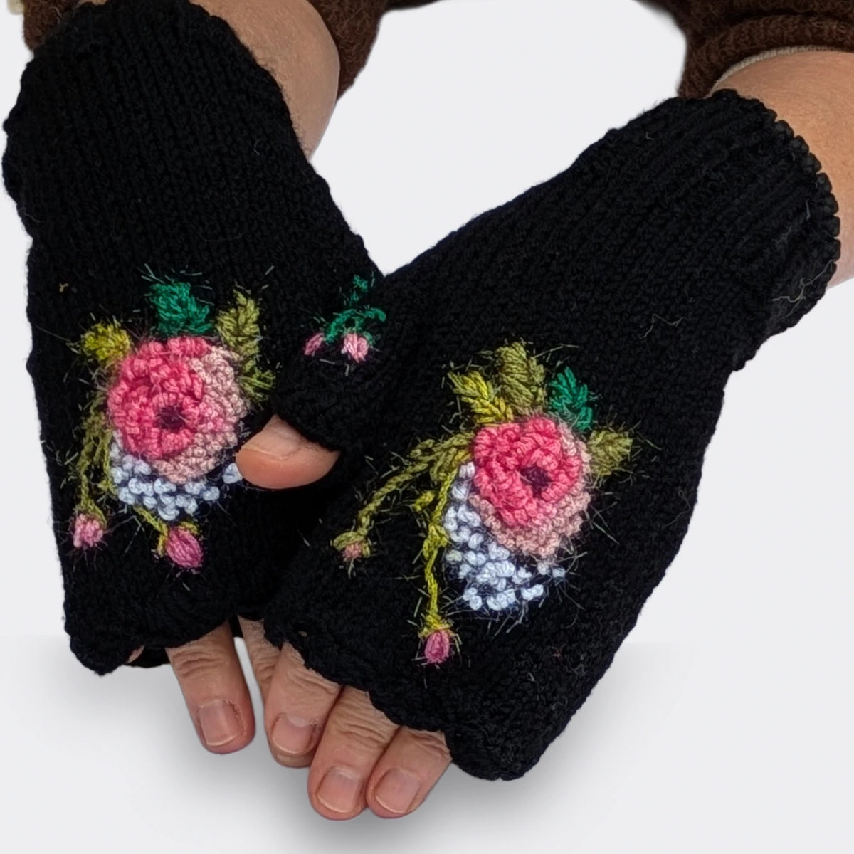 Mitaines noires tricotées main, portées sur des mains légèrement croisées. Une rose crochetée aux nuances de rose clair et de rose soutenu se détache parmi les petits points blancs et les tiges vert olive.