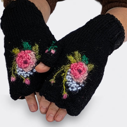 Mitaines noires tricotées main, portées sur des mains légèrement croisées. Une rose crochetée aux nuances de rose clair et de rose soutenu se détache parmi les petits points blancs et les tiges vert olive.