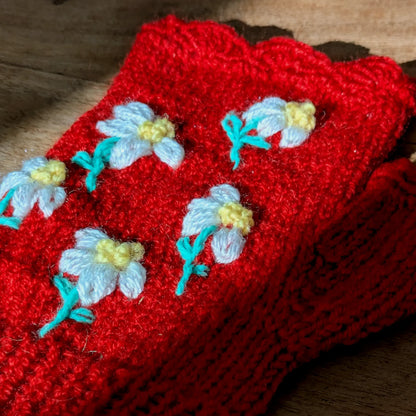 Vue supérieure d'une paire de mitaines rouges exposée à plat sur une table où passe un rai de lumière ensoleillé. De petites fleurs très mignonnes ornent le dessus.