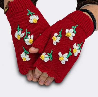 Mitaines rouges tricotées main portées sur des mains légèrement croisées. De petites fleurs blanches à cœur jaune et à tiges vertes sont réparties sur la maille rouge.
