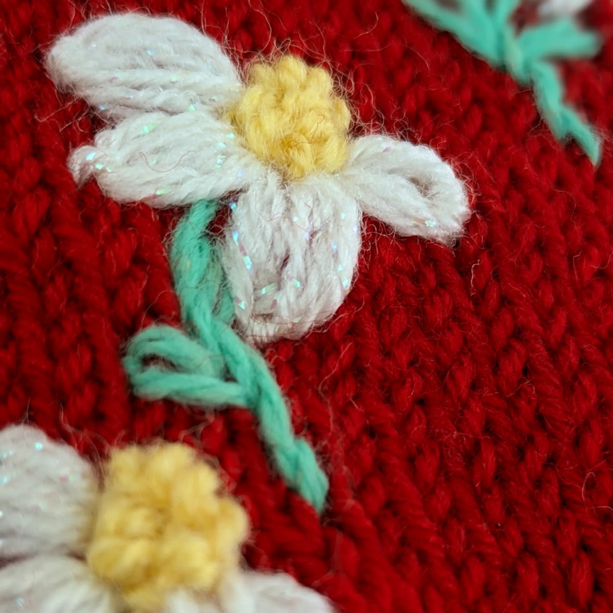 Vue en gros plan d'une des fleurettes brodée sur une paire de mitaines rouges. La petite fleur a un coeur jaune et des pétales blancs.