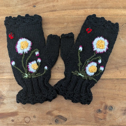 Paire de mitaines noires tricotées main, bord supérieur et inférieur festonné, pâquerettes et coccinelles brodées sur le dessus. Présentation à plat sur une table.