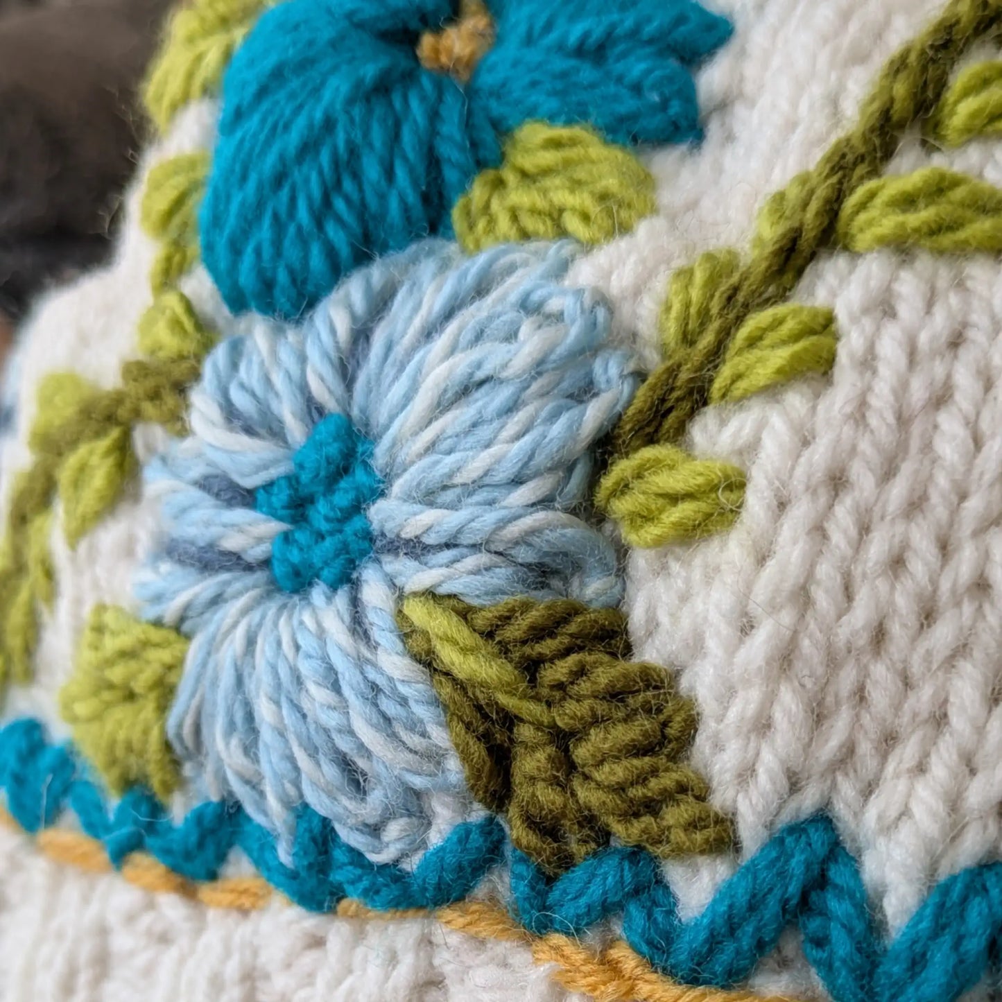 Détail des fleurs turquoise et des broderies végétales sur des mitaines tricotées main blanc cassé.