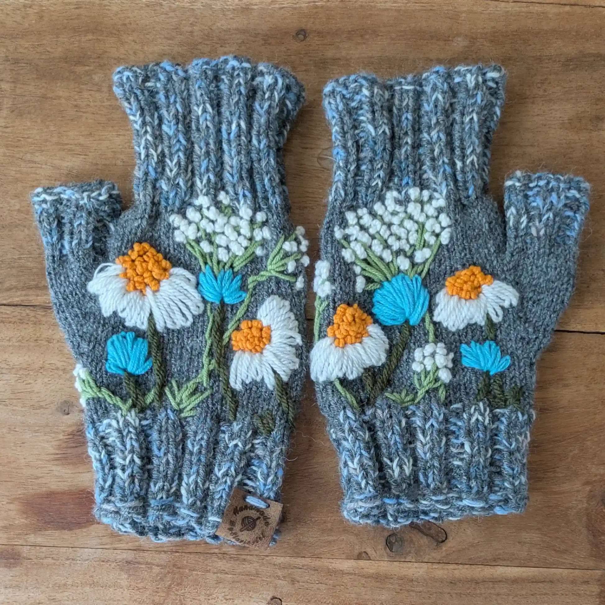 Mitaines grises tricotées main posées à plat, avec fleurs de camomille blanches et jaunes appliquées sur la maille.