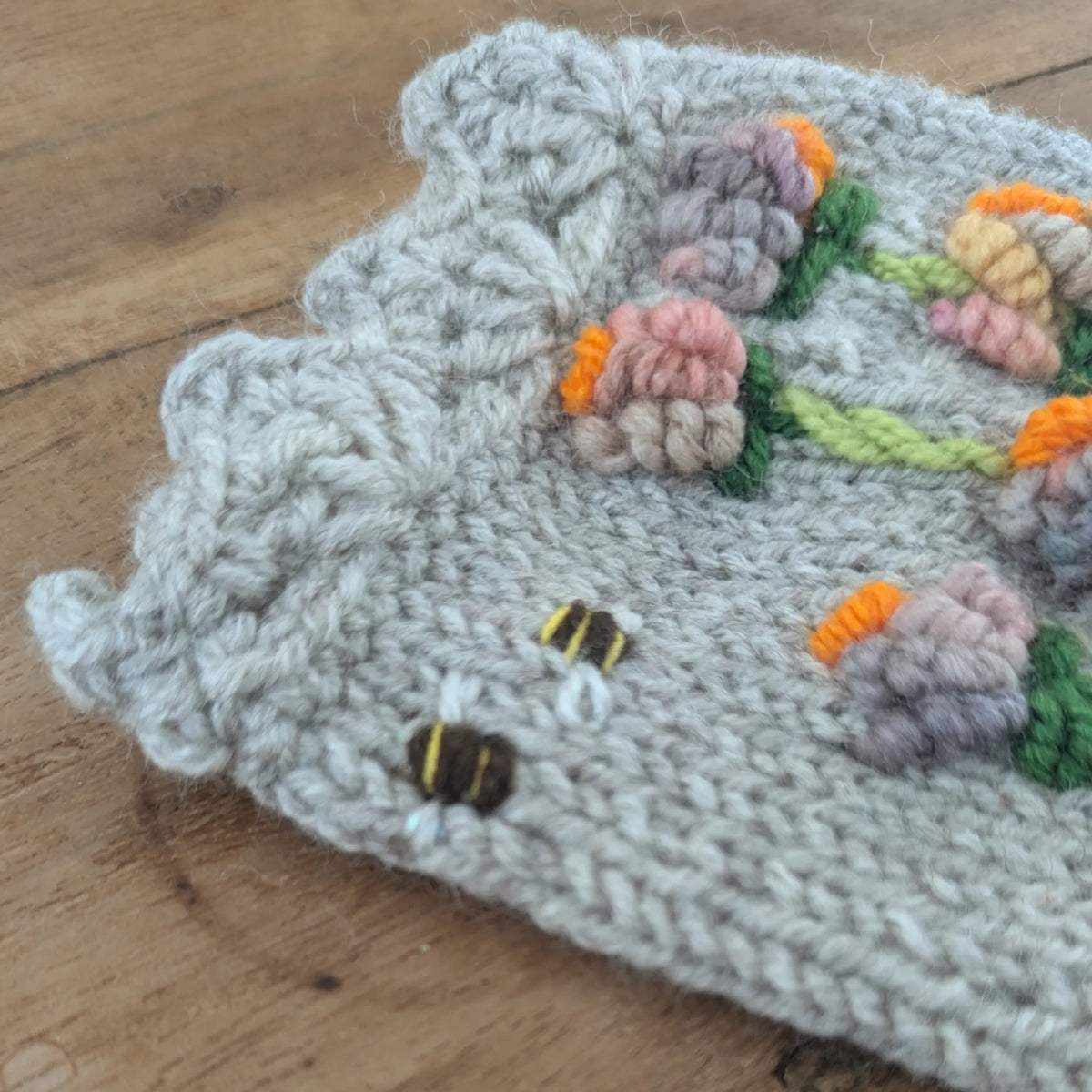 Gros plan sur la bordure festonnée de mitaines tricotées main en maille grise. Deux petites abeilles aux corps rayés de jaune et noir apparaissent à proximité des fleurs multicolores.