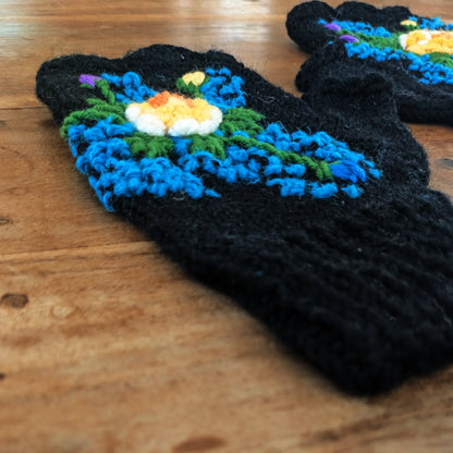 La photo montre une paire de mitaines noire tricotée main décorée d'une fleur centrale jaune et de petites fleurs bleues tout autour comme autant de points lumineux