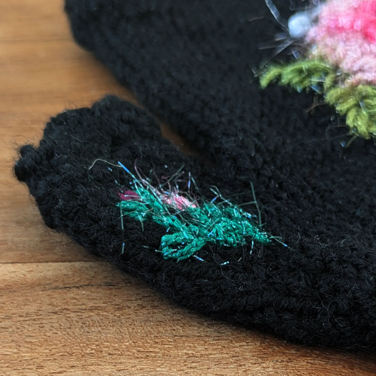 Détail du pouce d'une paire de mitaines noires artisanales. Une toute petite composition fleurie reprend le décor floral principale des mitaines.