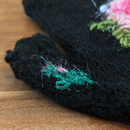 Détail du pouce d'une paire de mitaines noires artisanales. Une toute petite composition fleurie reprend le décor floral principale des mitaines.