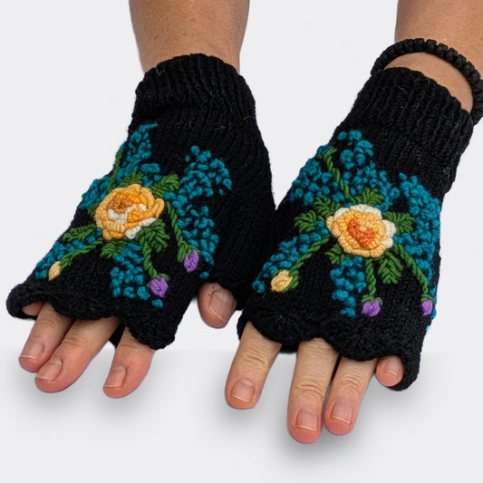 Mitaines artisanales noires portées sur des mains à plat, laissant apparaître le décor floral sur le dos des mains. Autour de la fleur centrale blanche et jaune, de petites touches bleues rayonnent parmi les feuilles vertes.