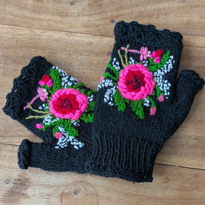 Mitaines noires tricotées main posées à plat, avec grande fleur rose appliquée en relief et broderies florales.