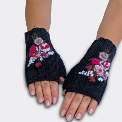 Vue à plat de mitaines artisanales bleu nuit. Les roses anciennes rose pâle et fuchsia sont appliquées en relief sur la maille côtelée. De petites fleurs blanches et des tiges vertes complètent le décor.