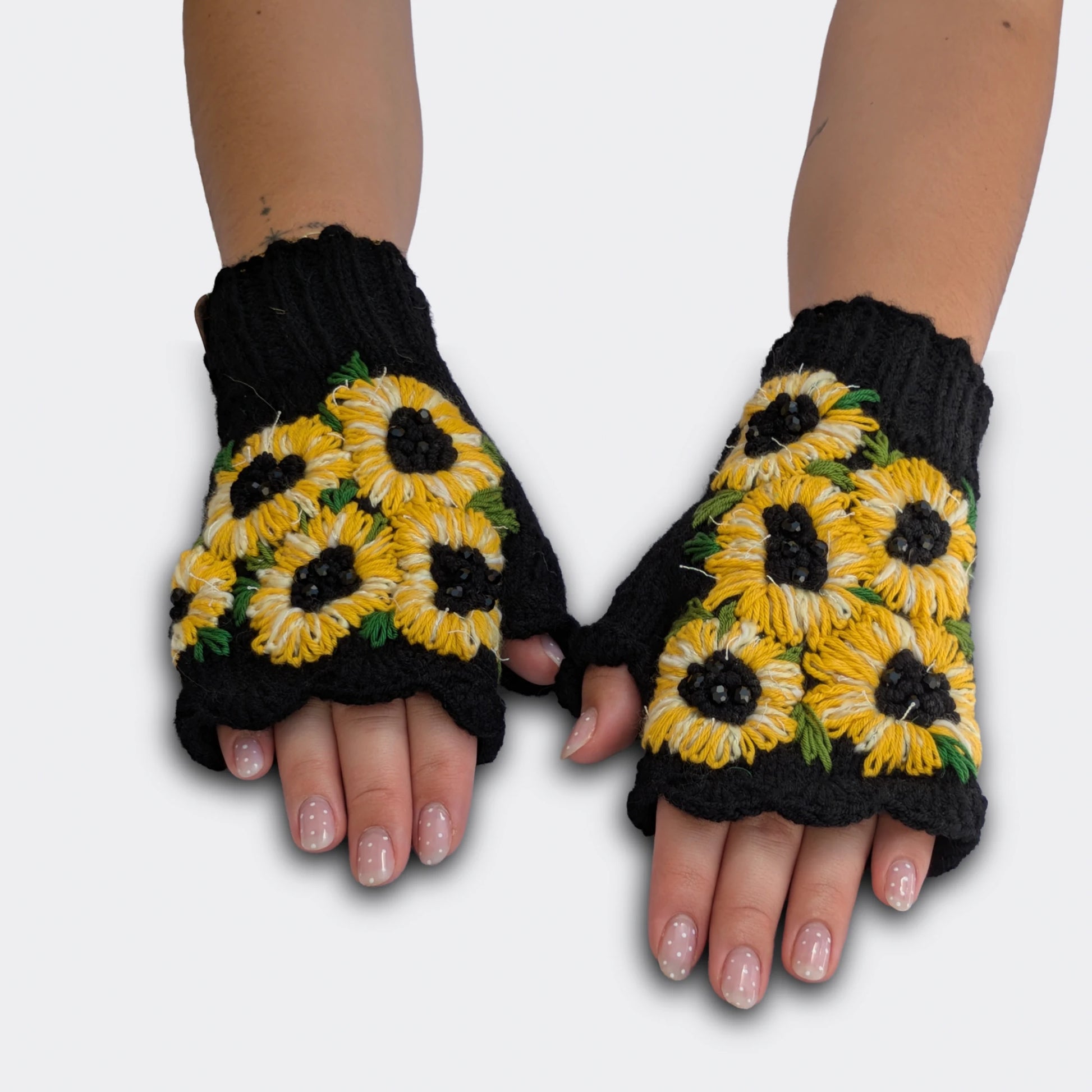 Mitaines tricotées main sur fond noir, portées à plat, avec tournesols en relief, cœurs noirs crochetés et feuillage vert.