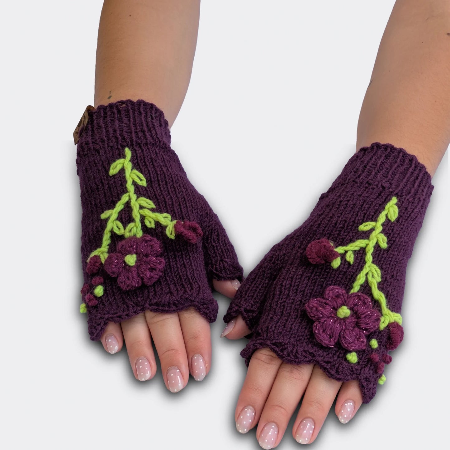Mitaines tricotées main sur fond violet, portées à plat, avec fleurs violettes appliquées et décor végétal vert clair.