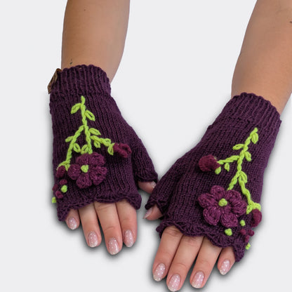 Mitaines tricotées main sur fond violet, portées à plat, avec fleurs violettes appliquées et décor végétal vert clair.