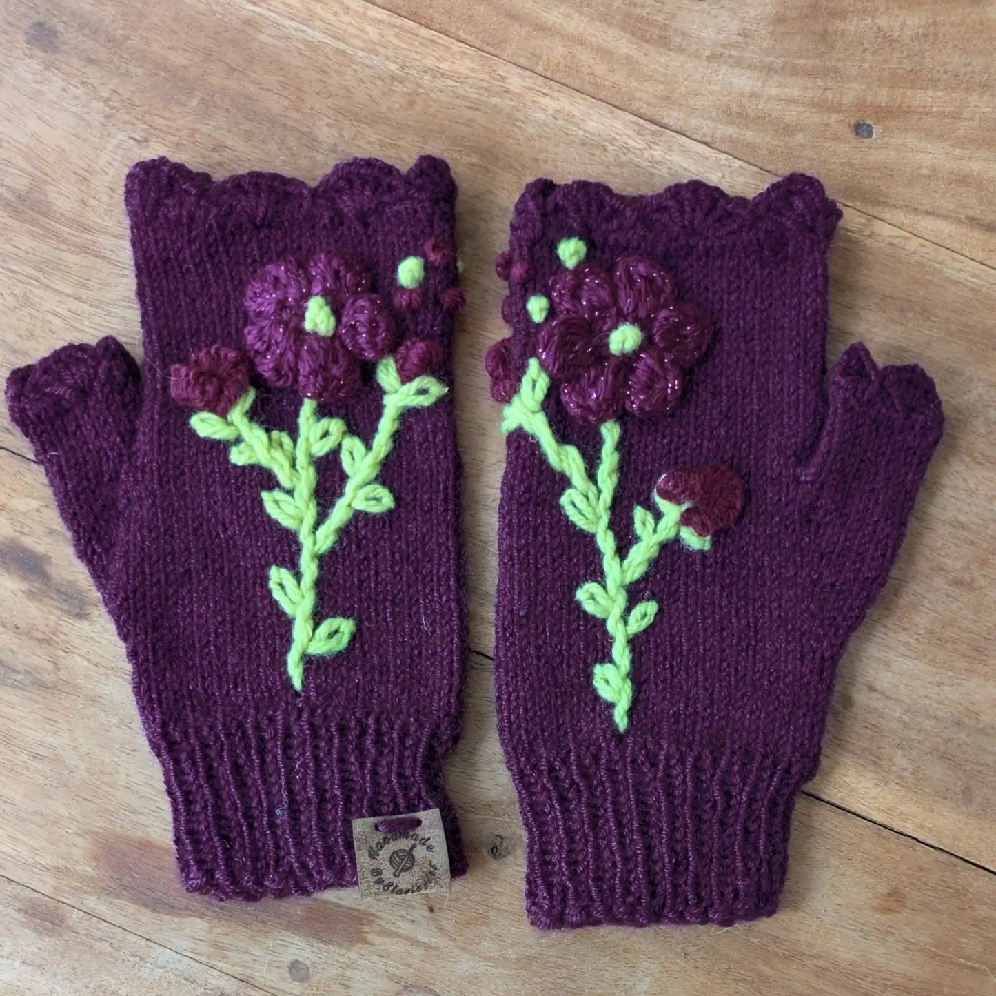 Mitaines violettes tricotées main posées à plat, avec tiges vert clair brodées et fleurs crochetées appliquées.