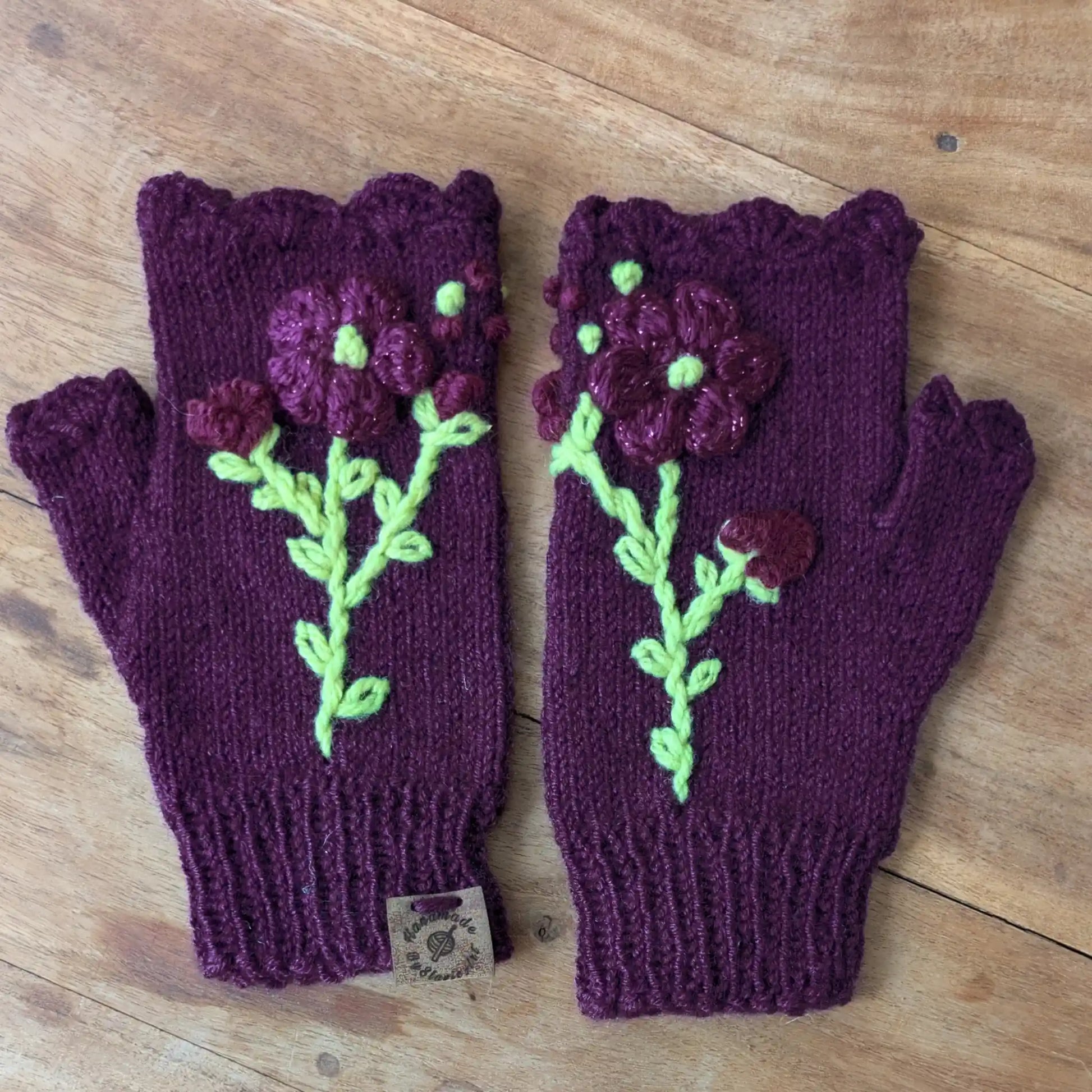 Mitaines violettes tricotées main posées à plat, avec tiges vert clair brodées et fleurs crochetées appliquées.