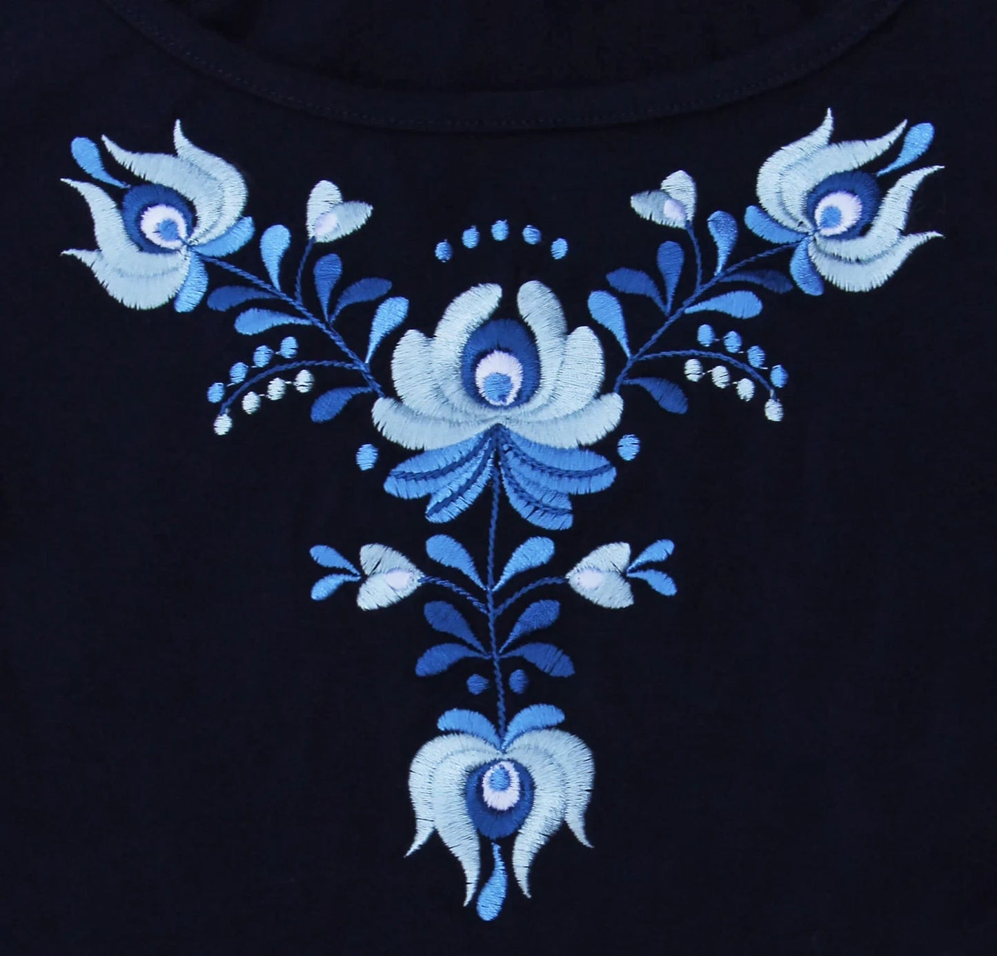 Gros plan de la broderie hongroise qui orne le t shirt bleu à manches courtes.  Vue de face des motifs, si les fleurs sont toutes en nuance de bleu leurs formes sont typiques des broderies Matyó.