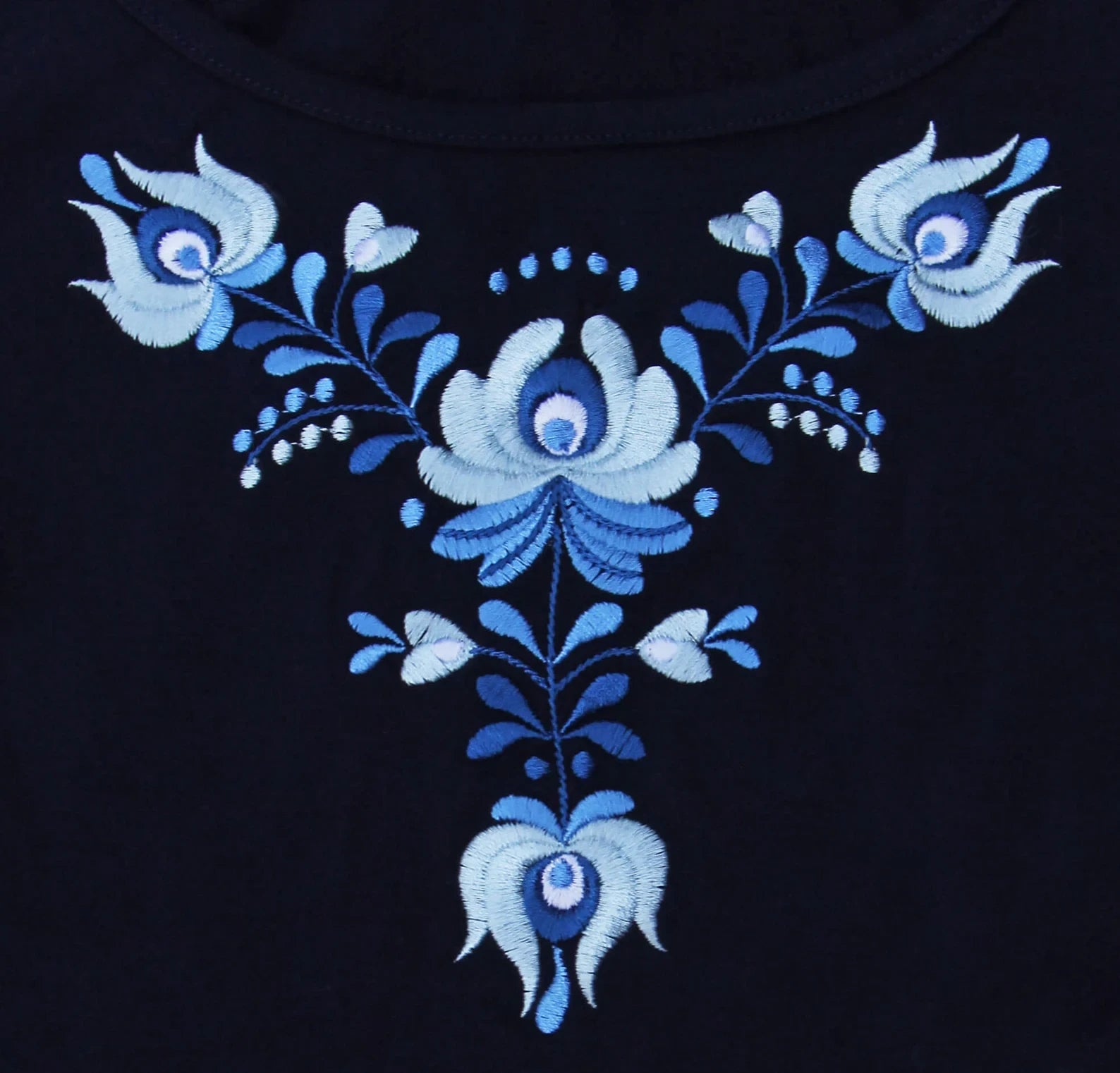 Gros plan de la broderie hongroise qui orne le t shirt bleu à manches courtes.  Vue de face des motifs, si les fleurs sont toutes en nuance de bleu leurs formes sont typiques des broderies Matyó.