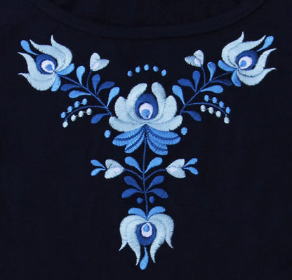 Gros plan de la broderie hongroise qui orne le t shirt bleu à manches courtes.  Vue de face des motifs, si les fleurs sont toutes en nuance de bleu leurs formes sont typiques des broderies Matyó.