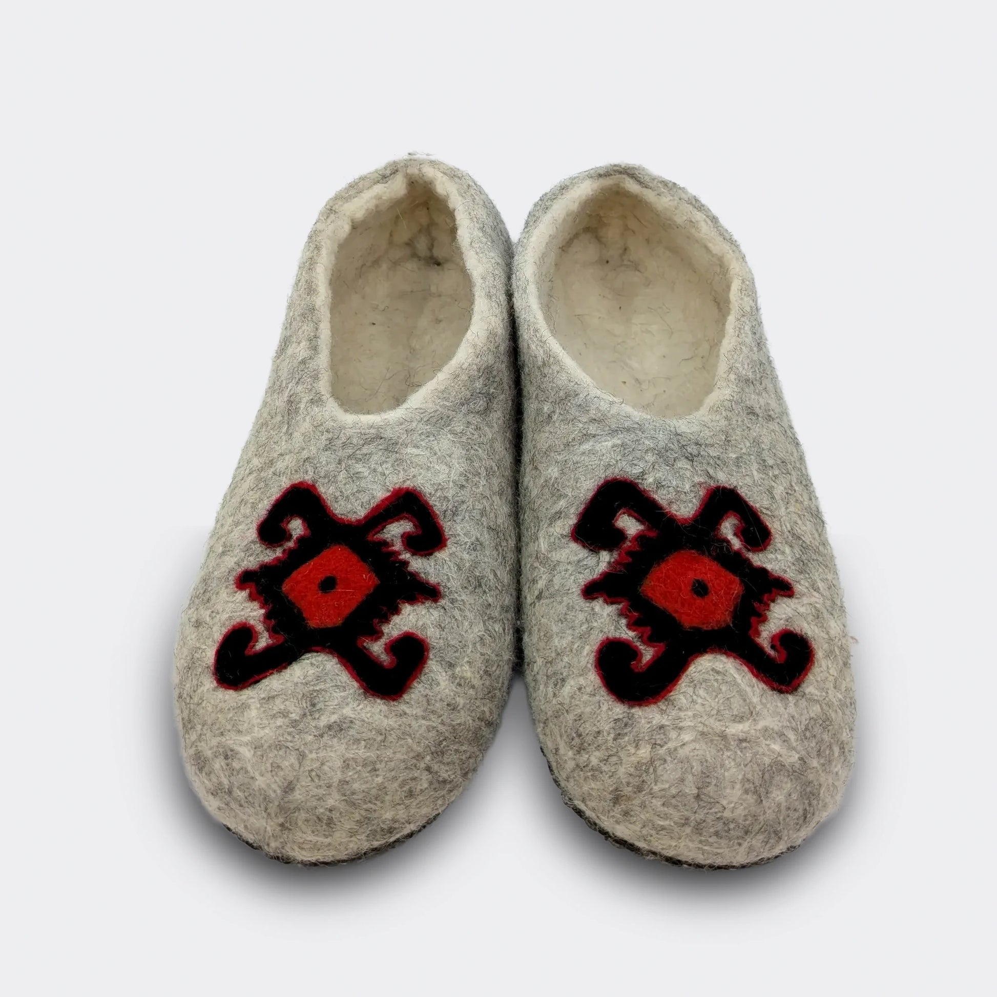 hausson en feutre gris vu de face, décoré du motif de la tortue des kilims de Pirot. Le dessin rouge et noir symbolise longévité et protection. La laine naturelle garde la mémoire d’un artisanat ancien. Vue de face.