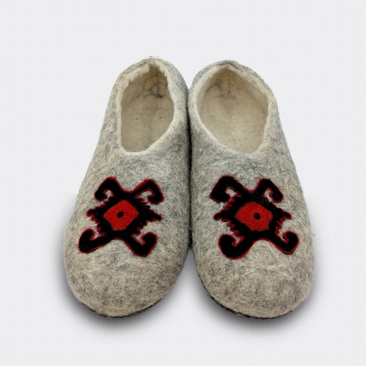 hausson en feutre gris vu de face, décoré du motif de la tortue des kilims de Pirot. Le dessin rouge et noir symbolise longévité et protection. La laine naturelle garde la mémoire d’un artisanat ancien. Vue de face.