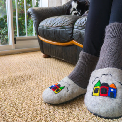 Une femme perte des pantoufles avec des maisons colorés dessinées dessus. Les pantoufles sont en feutre et réalisées à la main