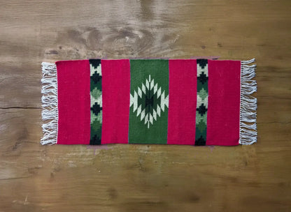 Petit tapis kilim disposé à plat avec des motifs traditionnels du Monténégro. Le kilim comprend un losange crénelé au centre dans une bande de coloris vert entouré par des bandes rouges.