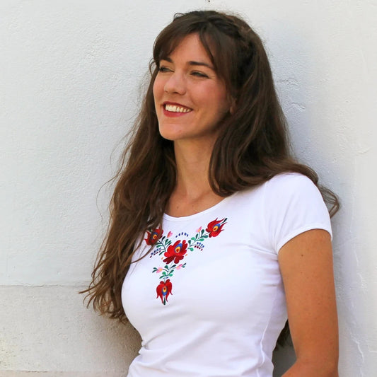T-shirt blanc à manches courtes brodé avec des fleurs rouges dans le plus pur style des broderies traditionnelles Matyó en Hongrie. La broderie est sous l'encolure et le col est rond. Photo de face, légèrement en biais.