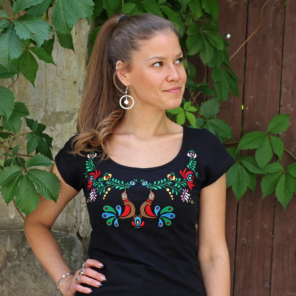 T-shirt noir à manches courtes avec une broderie caractéristique du style hongrois Matyó. Deux oiseaux colorés au centre se font face et se fondent parfaitement dans un décor floral magnifique aux teintes rouges, vertes et bleues. Vue rapprochée, coupée à la taille.